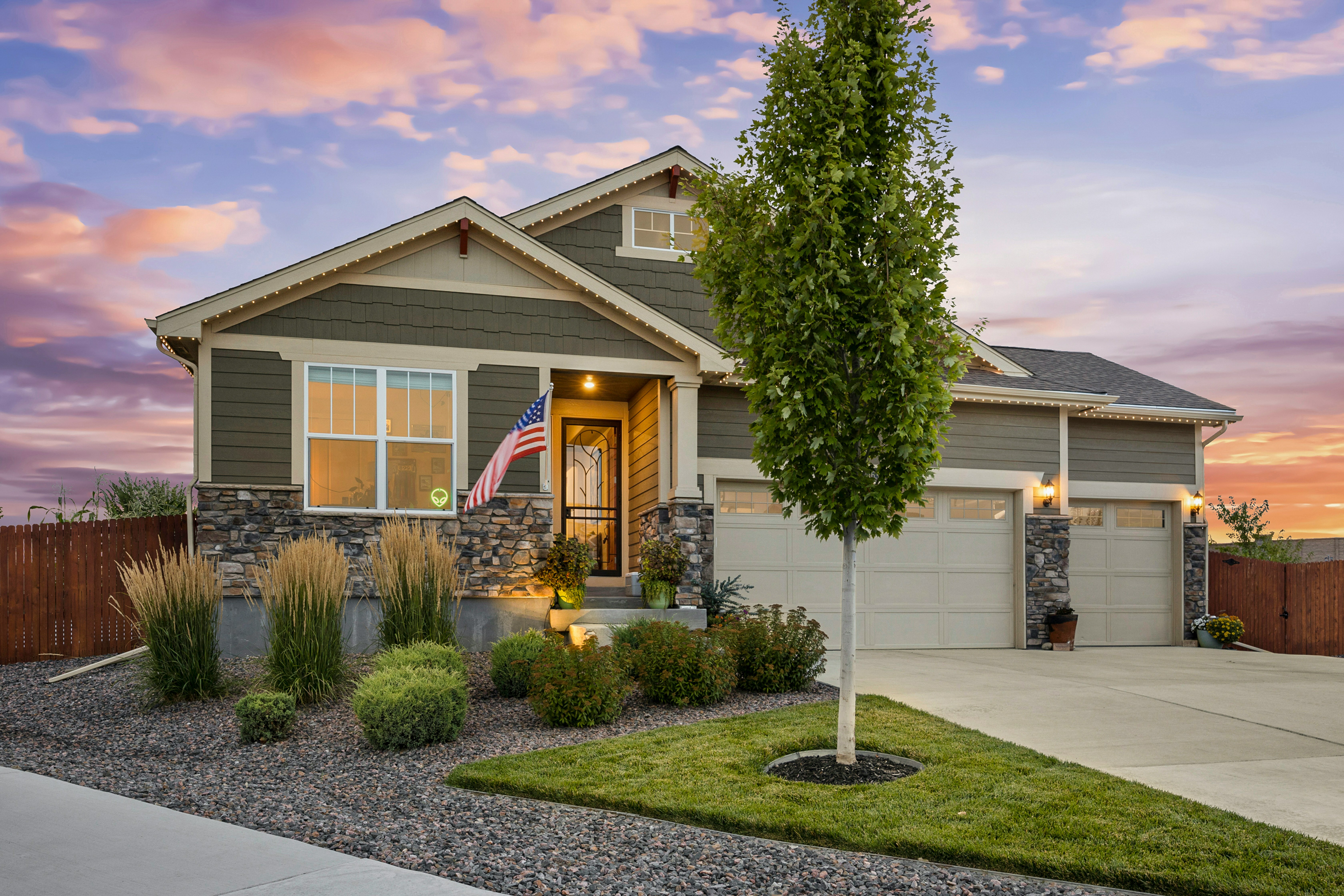 A tan home with stone accents has a paved driveway and stunning landscaping. A beautiful sunset is pictured in the background.